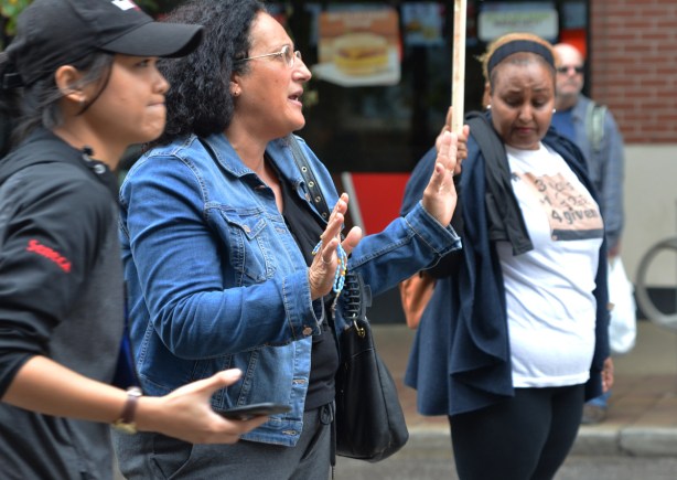 a middlw age woman in jean jacket and dark hair, holding a rosary in one hand, talks to counter protesters on the other side of a police line 