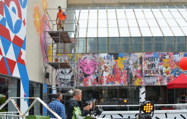 a man in an orange vest is up on scaffolding as he paints a mural, two other men are looking at finished murals