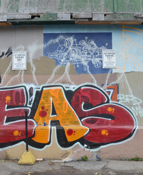 blue and white blueprint graffiti with large red and yellow text scrawl below it, on a wall in an alley