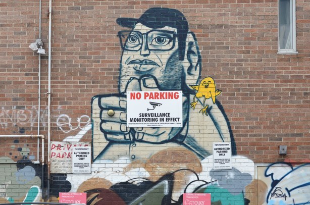 an old uber5000 painting on a wall of a man in black baseball cap and glasses with a little yellow birdie on his shoulder, a no parking sign on top of man's hands