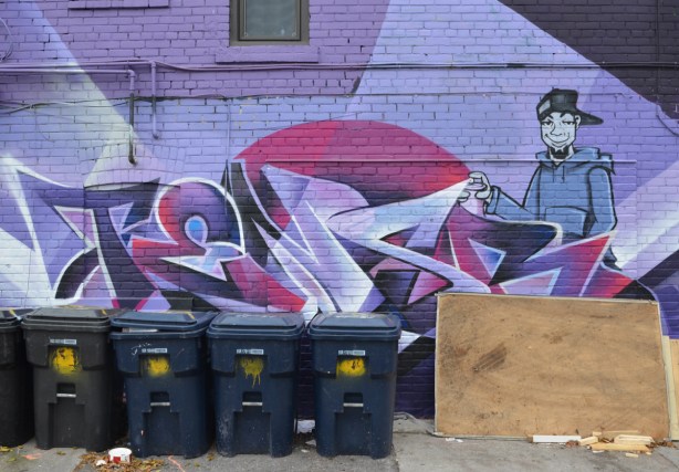 garbage bins, blue bins, in front of a purple wall with text street art and a painting of a young man with a can of spray paint
