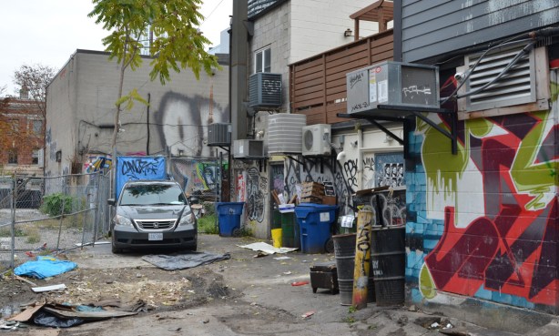 messy alley with car parked, trash bins, barrels, graffiti, air conditioners, back walls,