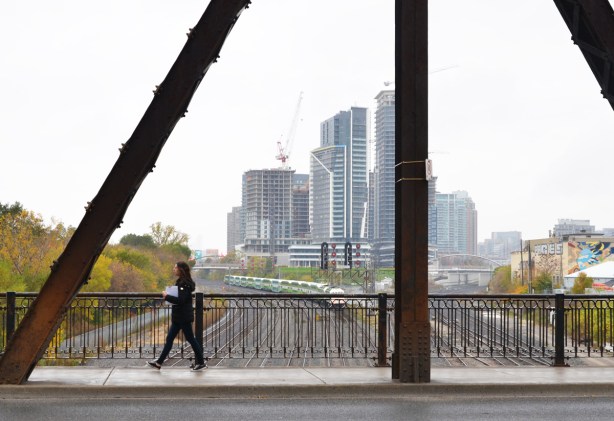 a woman walks over the bridge on Bathurst over the train tracks, GO train going westward in the background, as well as taller buildings to the west
