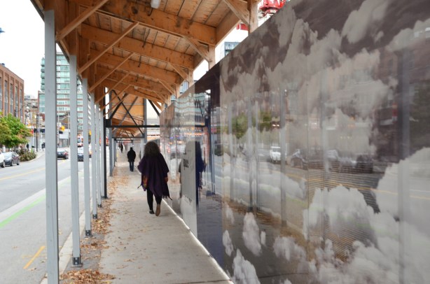 a woman walks past hoardings on Sherbourne street that are shiny and have pictures of clouds on them