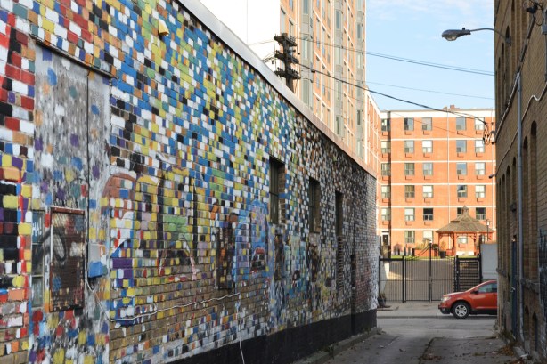 a brick wall where the bricks have been painted different colours like a giant mosaic, windows, and doors in that wall are also painted, in an alley,