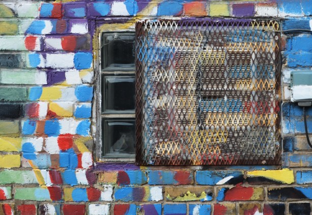 a brick wall where the bricks have been painted different colours like a giant mosaic, windows, and doors in that wall are also painted, in an alley,
