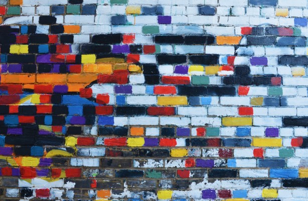 a brick wall where the bricks have been painted different colours like a giant mosaic, windows, and doors in that wall are also painted, in an alley,