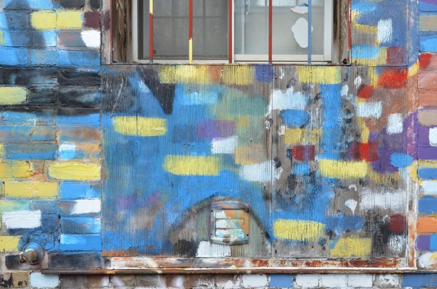 a brick wall where the bricks have been painted different colours like a giant mosaic, windows, and doors in that wall are also painted, in an alley, 