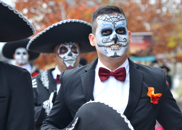 two men in white face day of the dead decorated, one with sombrero on and the other with the hat in his hand, wearing black suits, white shirts, and red bowties 
