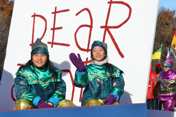 two girls dressed as Santa's elves sit on a parade float in front of a large white sign with red letters that says dear santa 