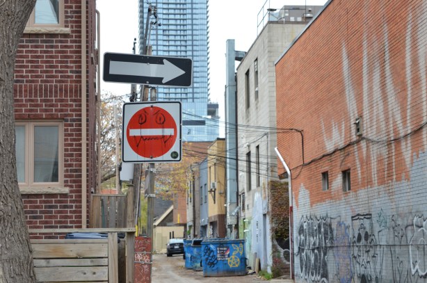 at the entrance to an alley, a red and white do not enter sign has been altered, a face has been drawn it in black sharpie