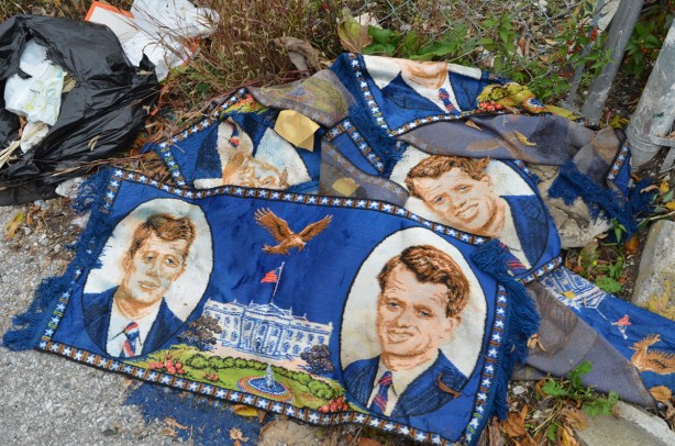 small blue carpets in a pile on the ground, about 4 of them, with pictures of John F Kennedy and Bobby Kennedy, a brown eagle, and some words
