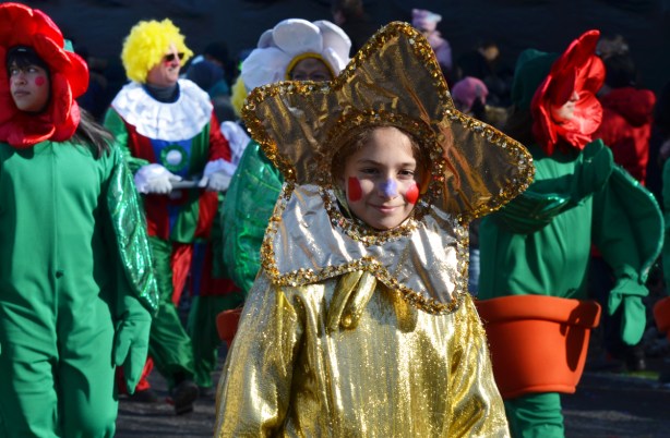 young woman walking in Santa Claus parade with large gold star shaped head-dress on, gold costume, other people dressed as plants in flower pots are nearby 
