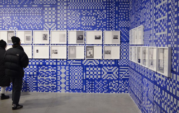 two people looking at framed pictures and pages of text on a wall that has been painted in blue and white squares