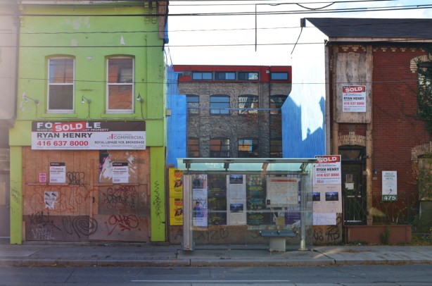 an empty bus shelter on Queen East, behind it is a vacant lot where a building had been destroyed by fire, to the left is a green building with bottom floor window boarded up and a sold sign on it. To the right is an old brick building