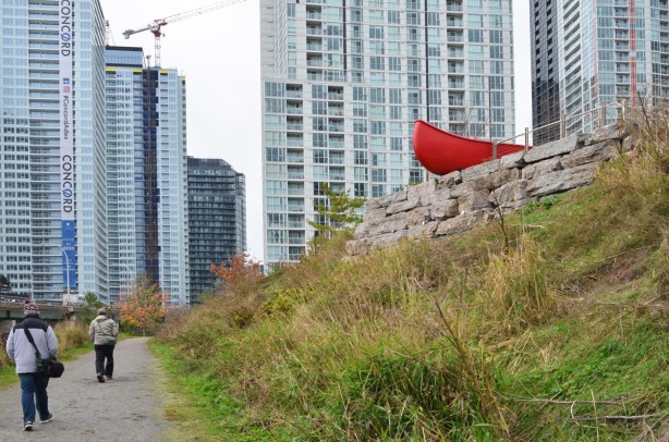 Douglas Coupland's red canoe at Canoe Landing Park sticks out of the edge of small hill, tall condo buildings in the background 