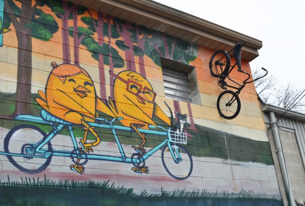 an UBer5000 mural of two yellow birdies on a tandem bike. An old bike is affixed to the wall beside the mural