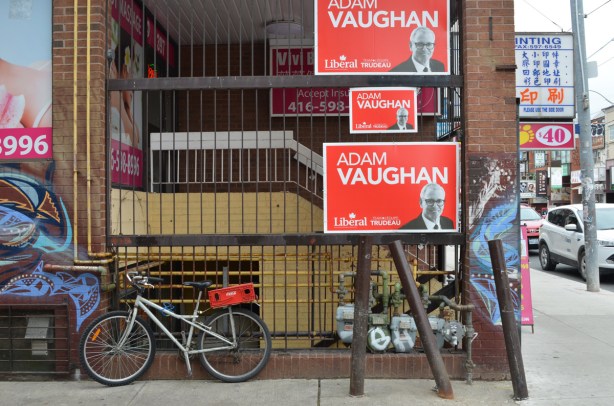 side of a building in Chinatown, stores and restaurant, bike parked there, also three large Adam Vaughan election signs.