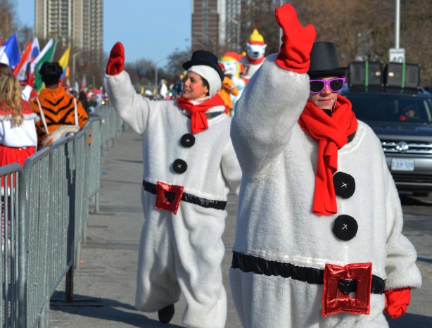 three women dressed as fat snowmen with black hats, black buttons, red scarves, and red belt buckles, waving to the crowd as they walk in the parade 
