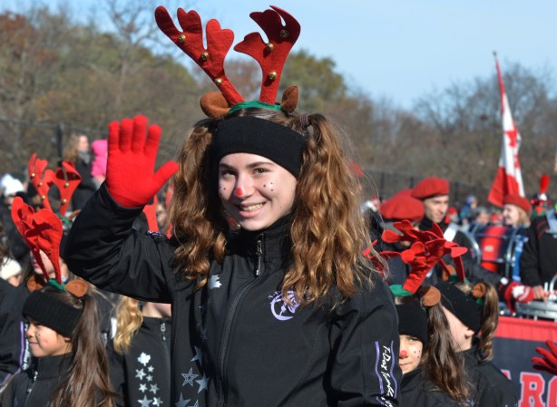 young woman in black jacket, red gloves, and large reindeer antlers, waving as she walks in Santa Claus parade 