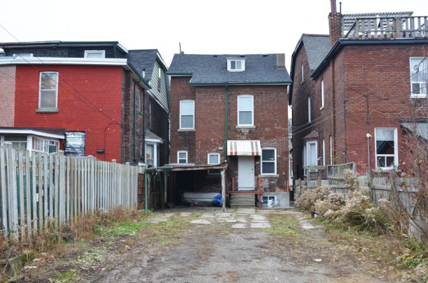 empty backyard of an older two storey building, with brick buildings on either side of it, seen from the laneway 