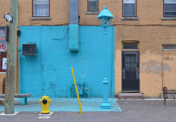 part of a lower storey of a building, as well as part of the sidewalk directly in front of it, are painted bright light blue