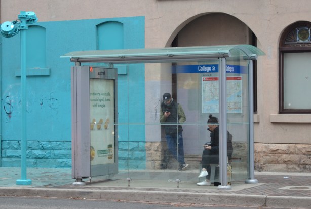 two people waiting in a TTC bus shelter, part of a lower storey of a building, as well as part of the sidewlk directly in front ot it, are painted bright light blue