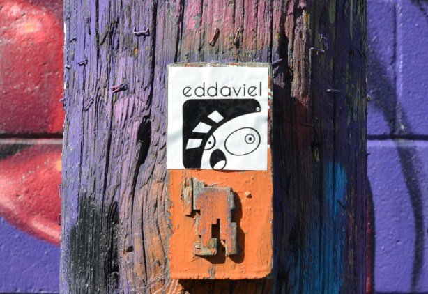 on a wood utility pole in graffiti alley, on top is a sticker with word eddoviel and on the bottom is the bottom part of an old orange stikman