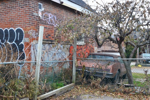 an old car is parked under a tree and beside a house with graffiti on it 