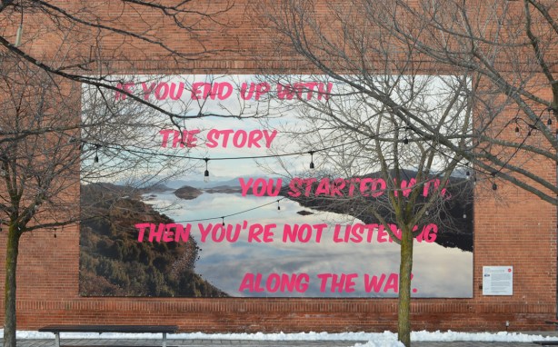 an art installation on the exterior wall of the Power Plant Contemporary Art Gallery, words in pink letters written over an river landscape scene. the words say If you end up with the story that you started with, then you're not listening along the way. 