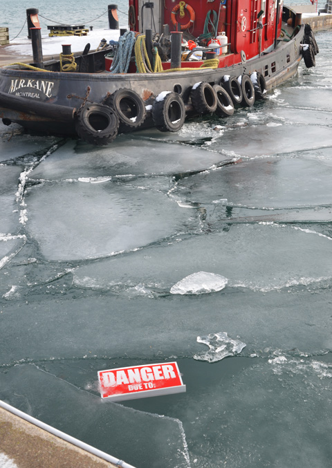 Toronto red tug boat in the water with brocken bits of ice in the water, also a red and white danger due to sign that has fallen onto the ice of Lake Ontario