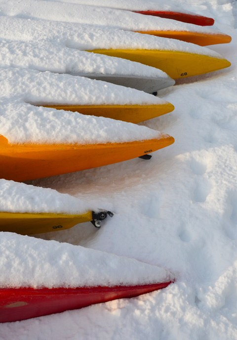 red, orange, and yellow kayaks on the ground for the winter, mostly covered with snow 