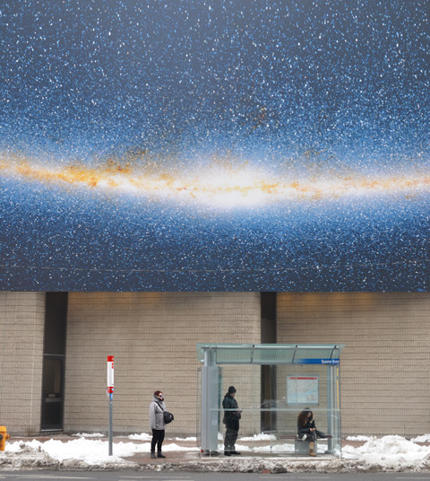 three people at a bus shelter waiting for a bis, two are standing and one is sitting. They are underneath a large photo of stars and the night sky 