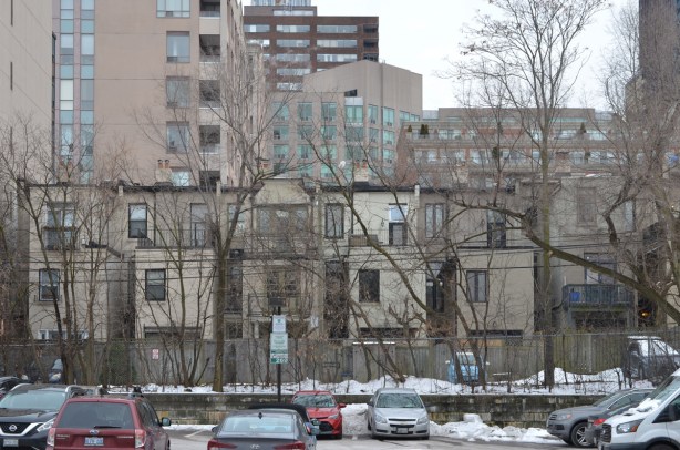 parking lot, with a row of backs of houses behind, with higher rise buildings in the background