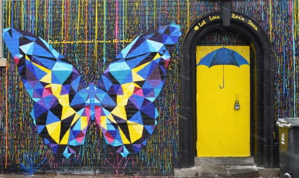 a multi coloured butterfly, mostly blue and yellow, made of geometric shapes, in a mural beside a yellow door with a blue umbrella painted on it