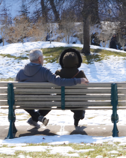 a couple sits on a bench, from behind, in winter clothes, snow on the ground in the park