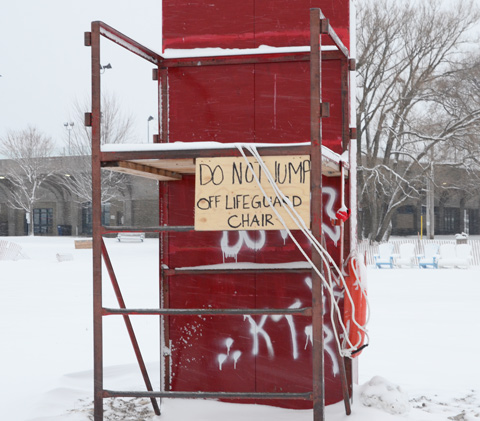 a metal frame lifeguard station, raised seat, with a red board against the back and a sign on the front that says do not jump off lifeguard chair 