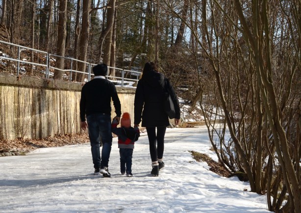a family of three, mom, dad, and toddler, walking on snowy path at Wilket Creek, with toddler in middle