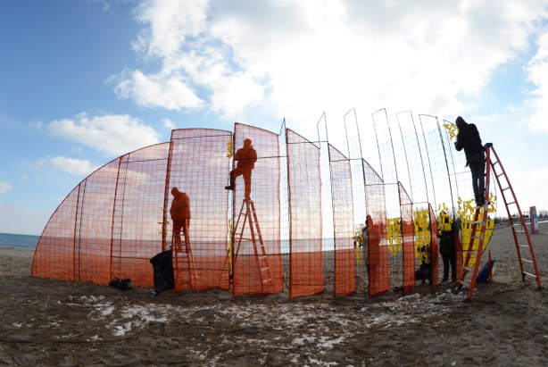 5 people working, two up on ladders, Lake Ontario in the background, tying yellow ribbons on an orange metal frame, finishing touches on an art installation called Mirage