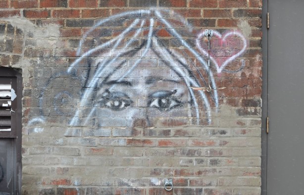 on a brick wall, a drawing of a woman's face with the eyes being the most prominent, a small red heart beside her face