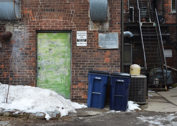 old beat up green door on the back of a brick building, lane, garbage bins there, also an old faded sign that says trespassers will be prosecuted, metal stairs leading up to upper storeys 