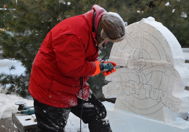 a man in a bright red winter jacket and brown hat and orange mitts is using a power drill to cut a picture of bugs bunny and the words looney tunes from a block of ice
