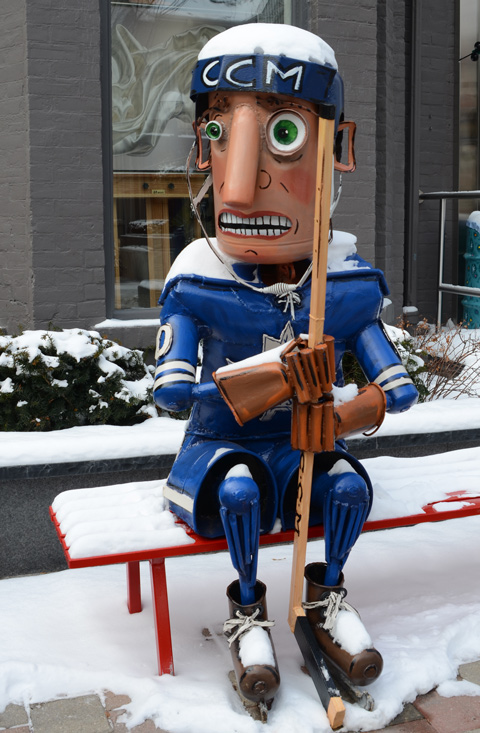 metal sculpture of a Toronto Maple Leaf hockey player sitting on a bench outside, holding his hockey stick and looking frightened, by Patrick Amiot