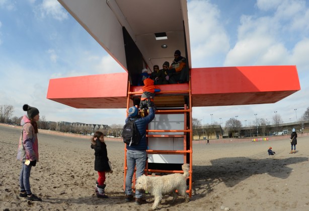 a father lifts up a young boy in a red winter coat so he can sit inside an art installation over a lifeguard station at the beach 
