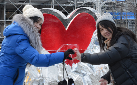 two women make a heart symbol with their hands in front of a red heart frozen inside an ice sculpture