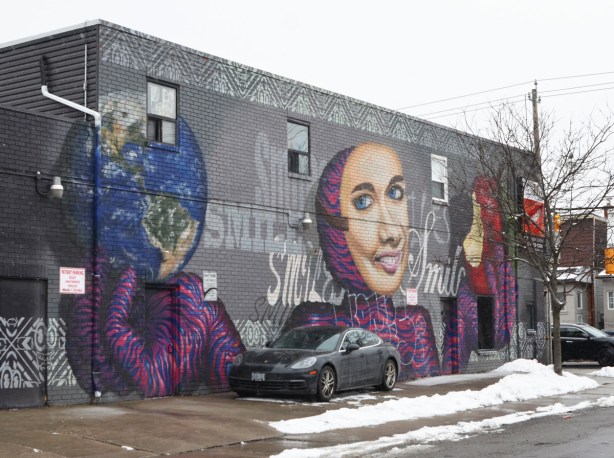 long mural by spud bomb of a woman smiling, holding a red apple with a bite out of it in one hand and a globe in the other hand. She is wearing a red and purple striped close fitting outfit over her arms and head. On the side of a dental office. The word smile is written many times in different fonts