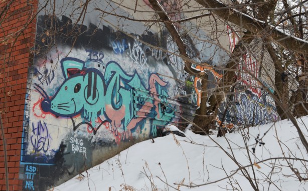graffiti on the side of a building by a small hill and some trees. The hill is part of the embannkment for the railway tracks