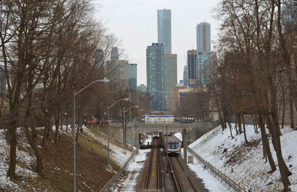looking down the TTC subway tracks from just north of Rosedale station, highrises of downtown in the background, trees beside the tracks, 2 subway cars, one going north and the other south 