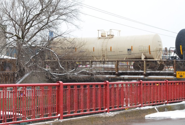 a red railing separates a parking lot from a hill, in the background a white tanker railway car is passing over a bridge