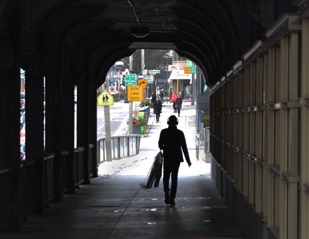 a man walks under a bridge, has headphones on and is carrying dry cleaning in a plastic cover 